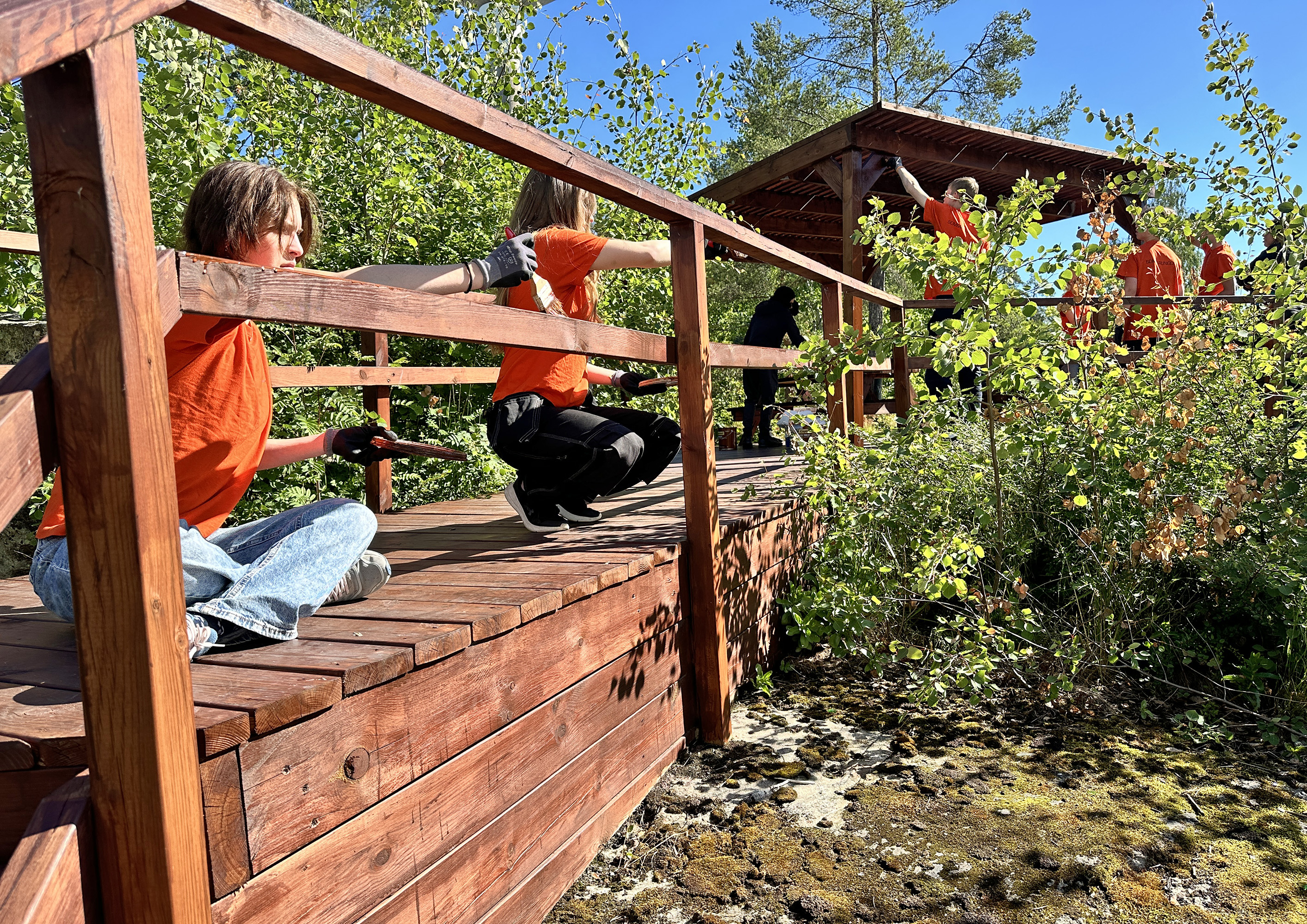 En grupp ungdomar i orange t-shirts oljar in en träkonstruktion utomhus.